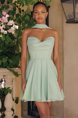 A woman modeling a strapless mint green sweetheart mini dress with a pleated skirt, standing in front of a beige wall with green foliage and pink flowers.