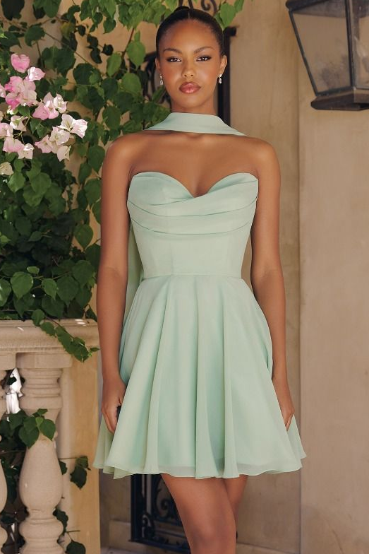 A woman modeling a strapless mint green sweetheart mini dress with a pleated skirt, standing in front of a beige wall with green foliage and pink flowers.